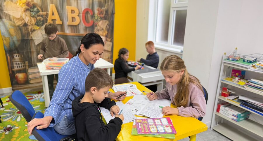 Educatoarea ajută copiii la teme, la masa galbenă din sala de clasă