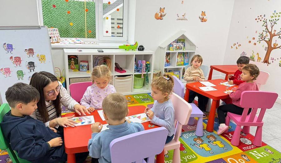 Educatoare care lucrează cu copiii pe fișe, la măsuțele mini-grădiniței